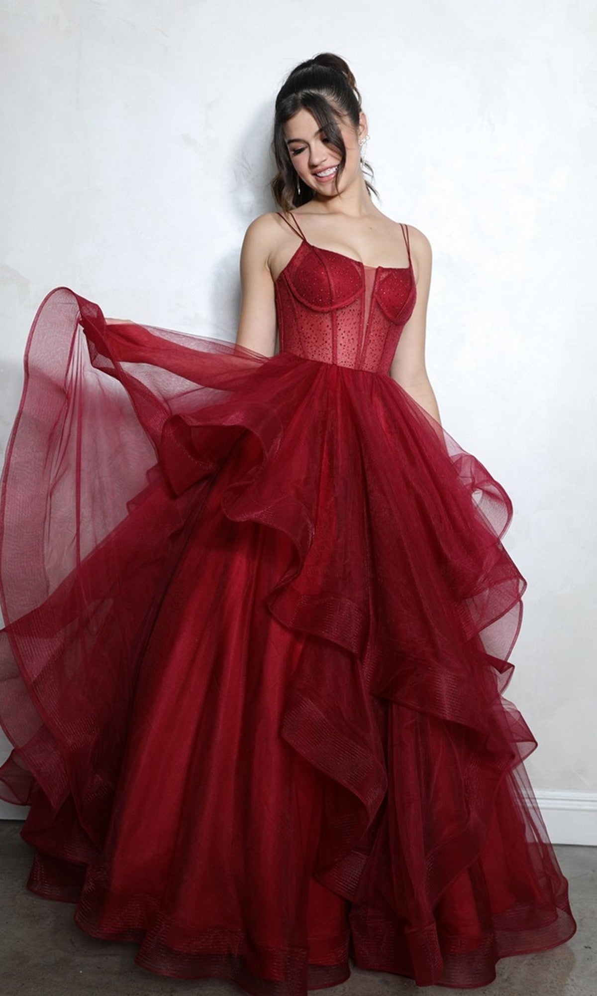 A woman stands indoors wearing the Eureka 9950 Long Prom Dress, a deep red gown with spaghetti straps, a voluminous ruffled tulle skirt, smiling as she holds out one side and looks down.